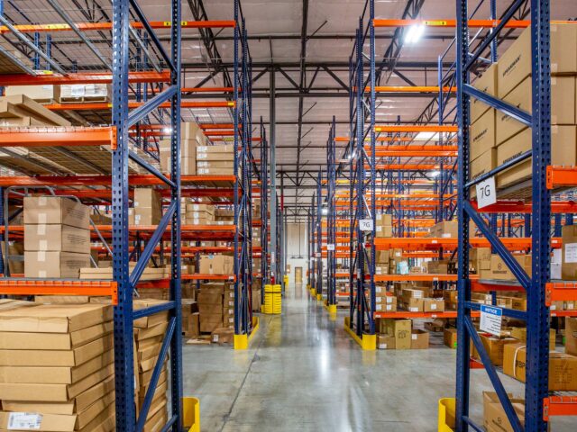 Warehouse aisle with tall blue and orange pallet racks stocked with boxes and HVAC parts.